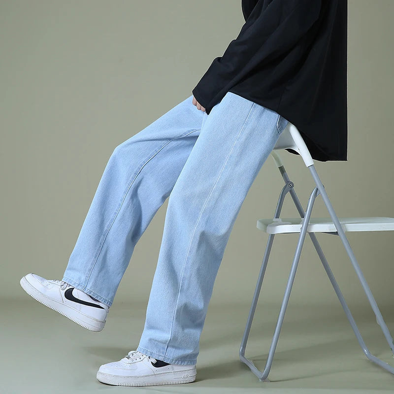 Man wearing light blue relaxed-fit wide-leg denim jeans with black top and white Nike sneakers, seated on white chair.