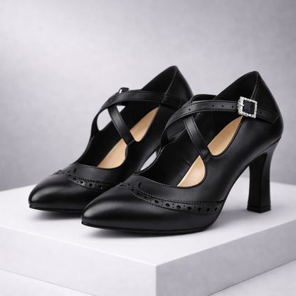 Black leather block heel pumps with cross straps, crystal buckle, and brogue detailing displayed on white platform.