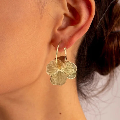 Gold flower hoop earrings worn on woman's ear, featuring textured floral pendant in brushed gold finish