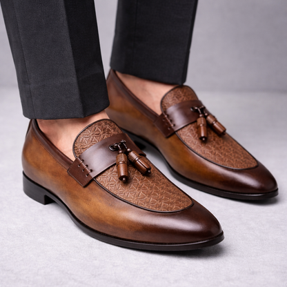 Men's brown leather tassel loafer dress shoes with embossed vamp detail, worn with dark charcoal trousers.
