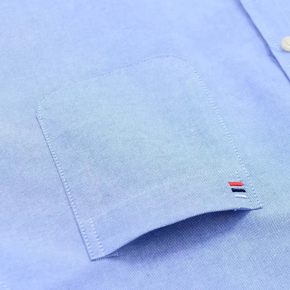 Light blue men's cotton button-down shirt close-up showing chest pocket, cuff, and red, navy, white logo detail.