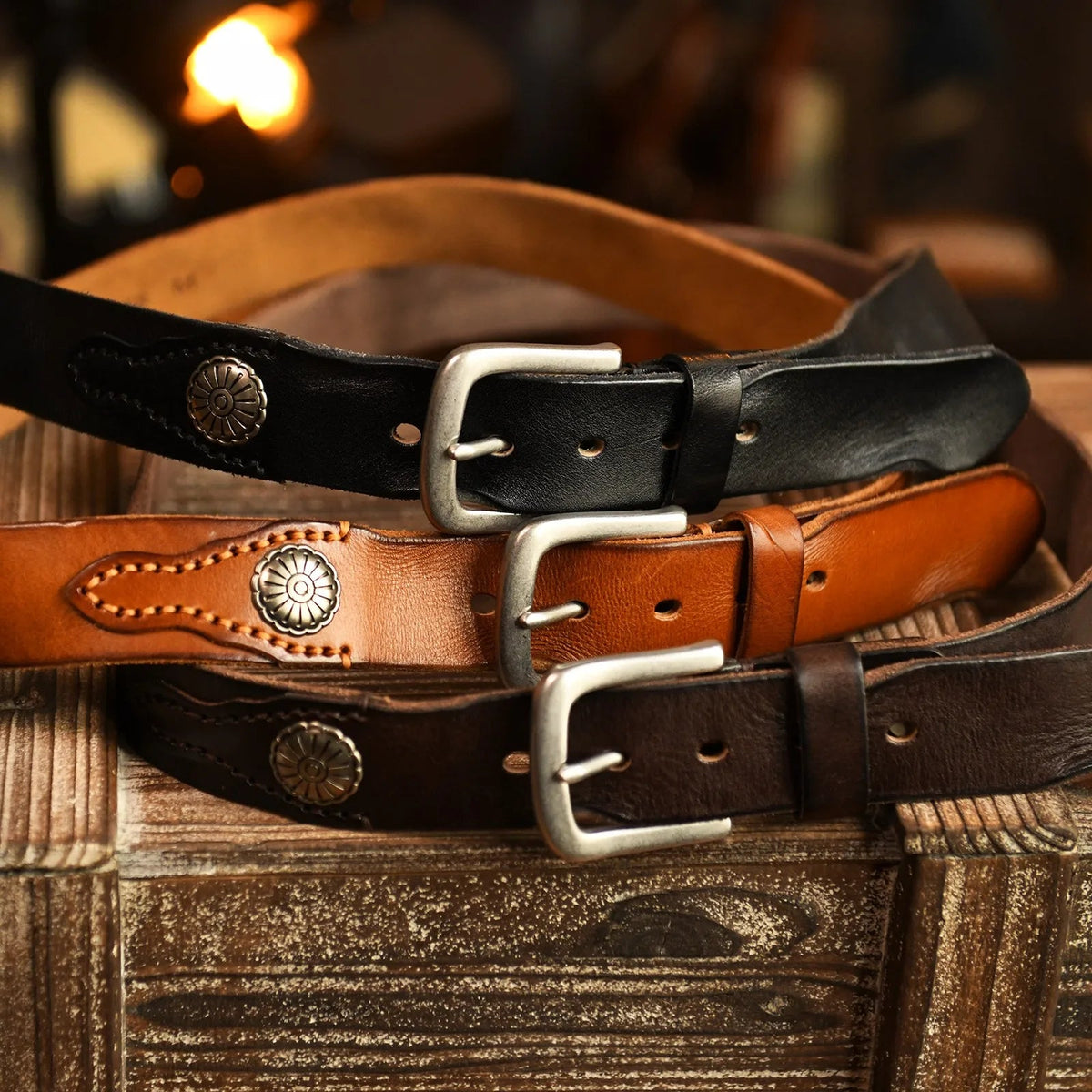 Three men's leather belts in black, tan, and brown with silver buckles and hand-stitched metal conchos on rustic wood.