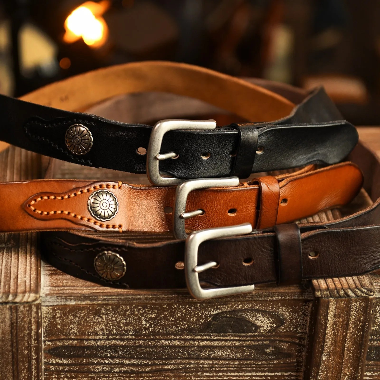 Three men's leather belts in black, tan, and brown with silver buckles and hand-stitched metal conchos on rustic wood.