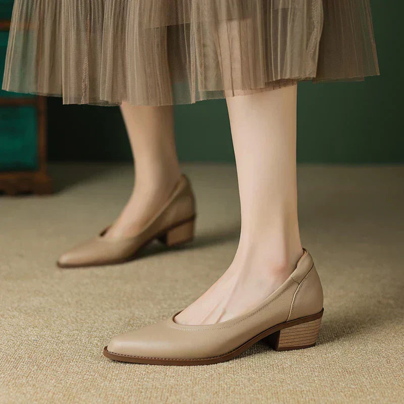 Women's beige leather pointed-toe pumps with block mid-heel, styled with a flowy mesh midi skirt on a neutral floor.