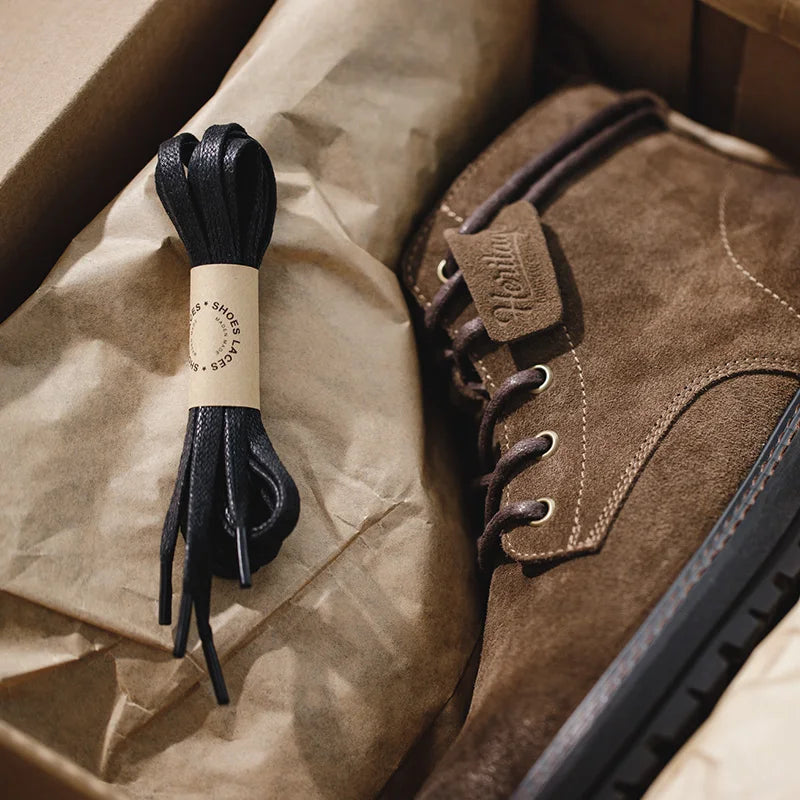 Brown suede men's lace-up ankle boots in open box with extra black waxed shoelaces on kraft paper packaging.