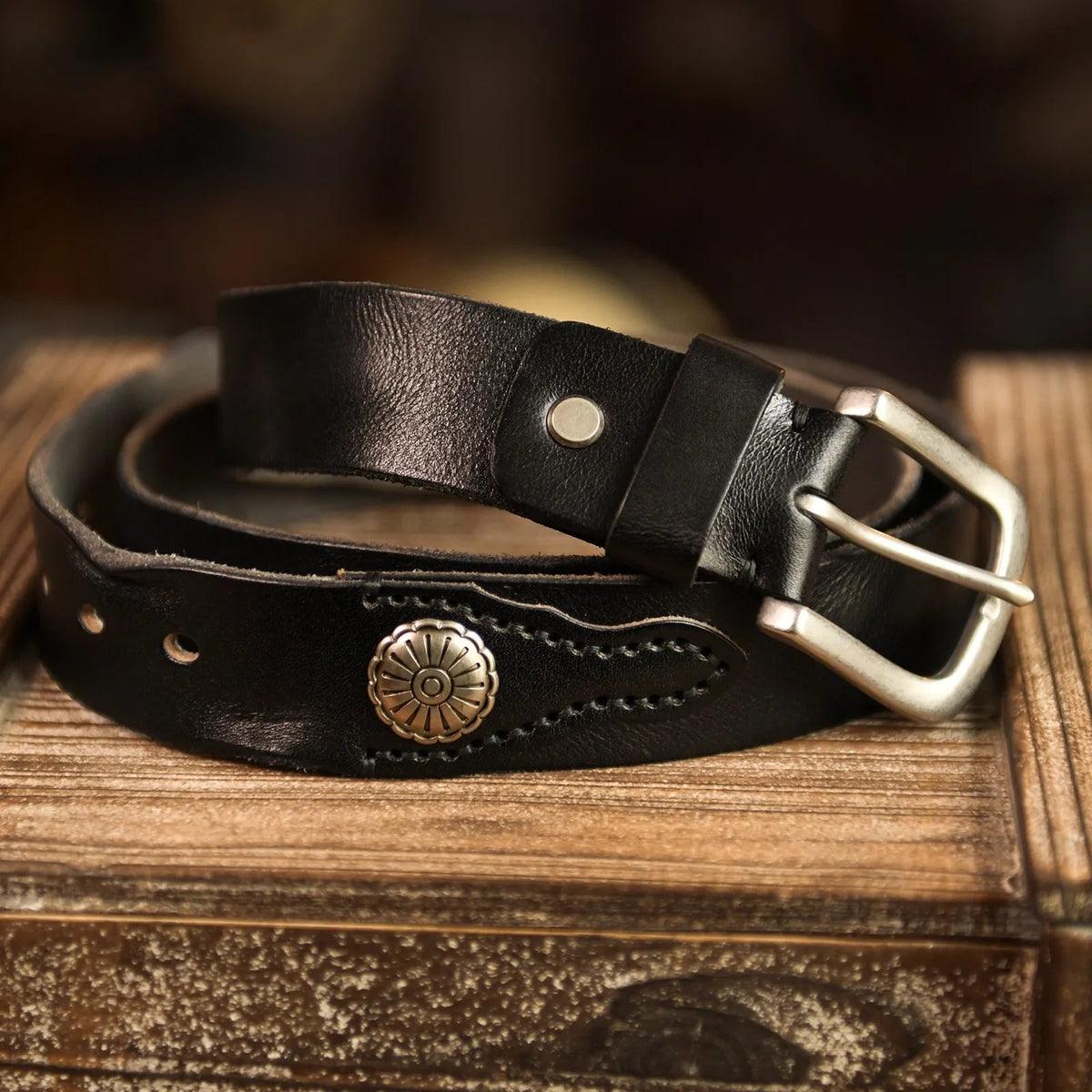Black leather men's belt with silver buckle, hand-stitched detailing, and decorative metal concho, coiled on rustic wood surface.