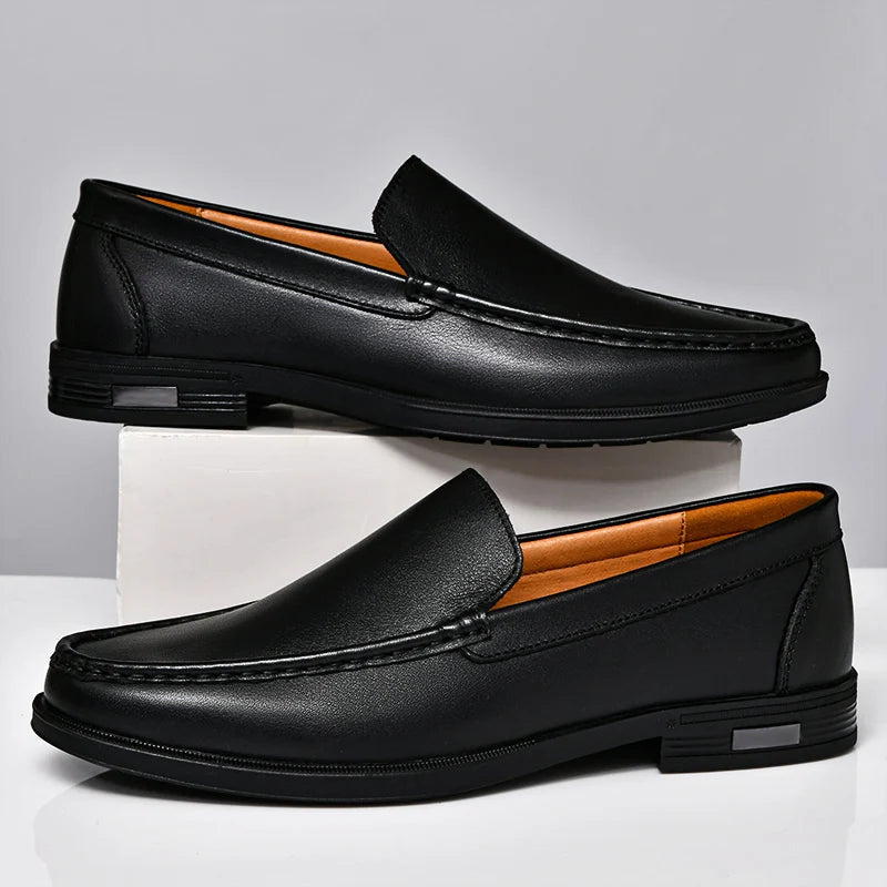 Black men's leather slip-on loafers with contrast stitching, tan interior lining, and low heel, displayed on white platform