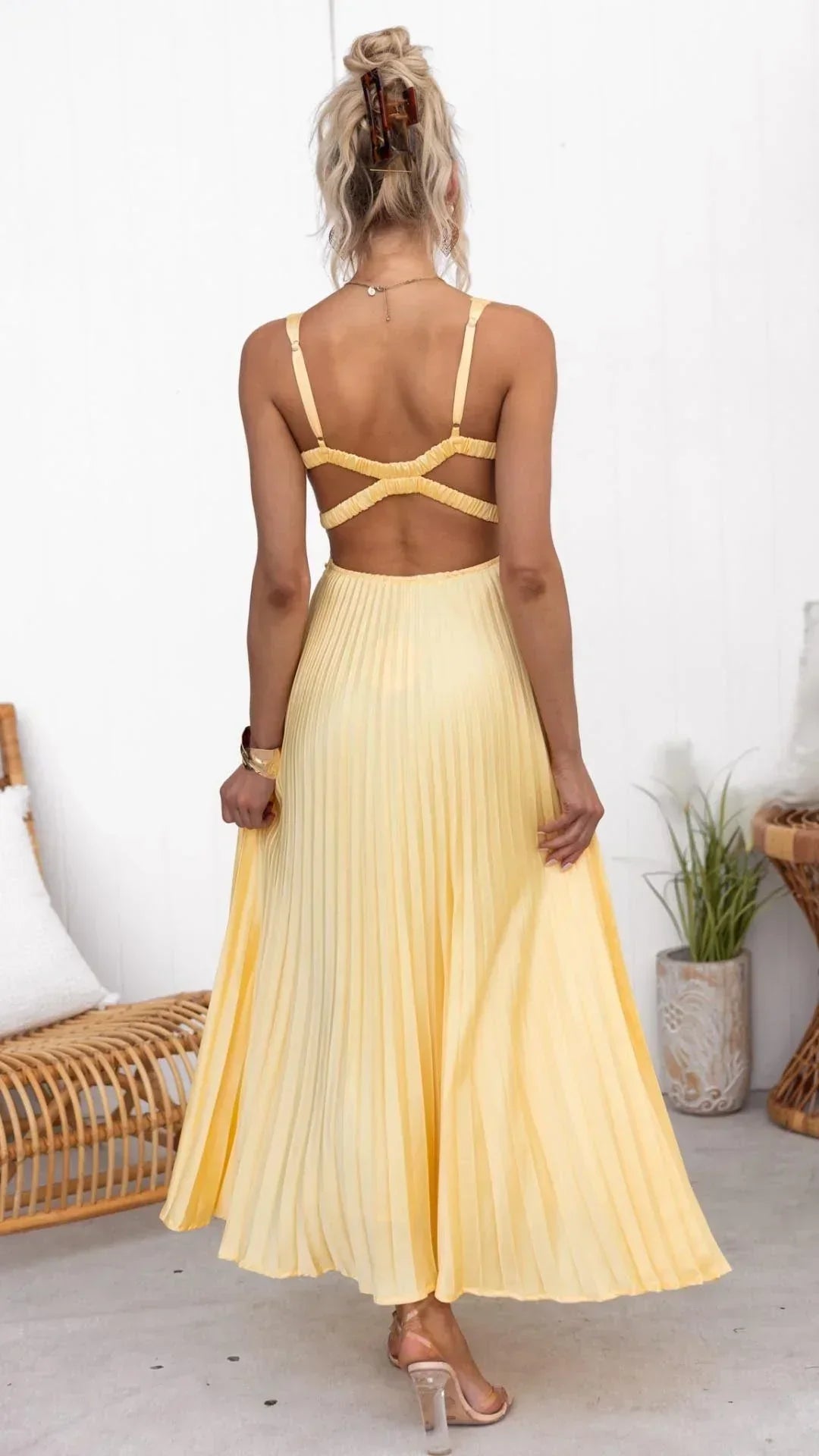 Yellow pleated maxi dress with open cross-back straps, spaghetti straps, and flowy skirt, worn with clear heeled sandals.