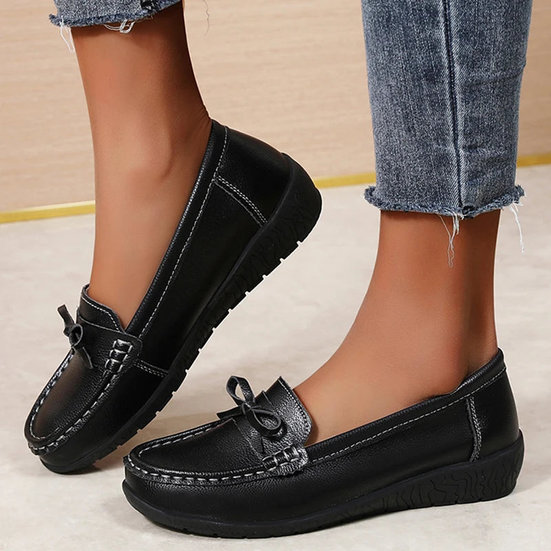 Black leather slip-on loafers with bow detail and cushioned sole, shown with distressed jeans on a woman's feet.
