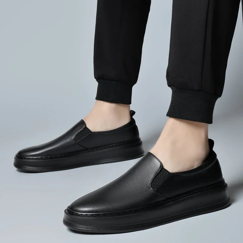 Men's black leather slip-on loafer sneakers worn with black jogger pants on gray background