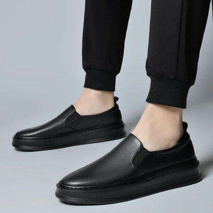 Men's black leather slip-on loafer sneakers worn with black jogger pants on gray background