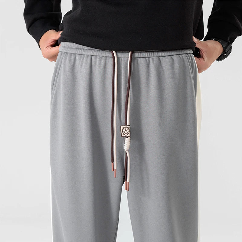 Men's gray wide-leg sweatpants with white side stripes, elastic drawstring waist, and smiley face charm detail.