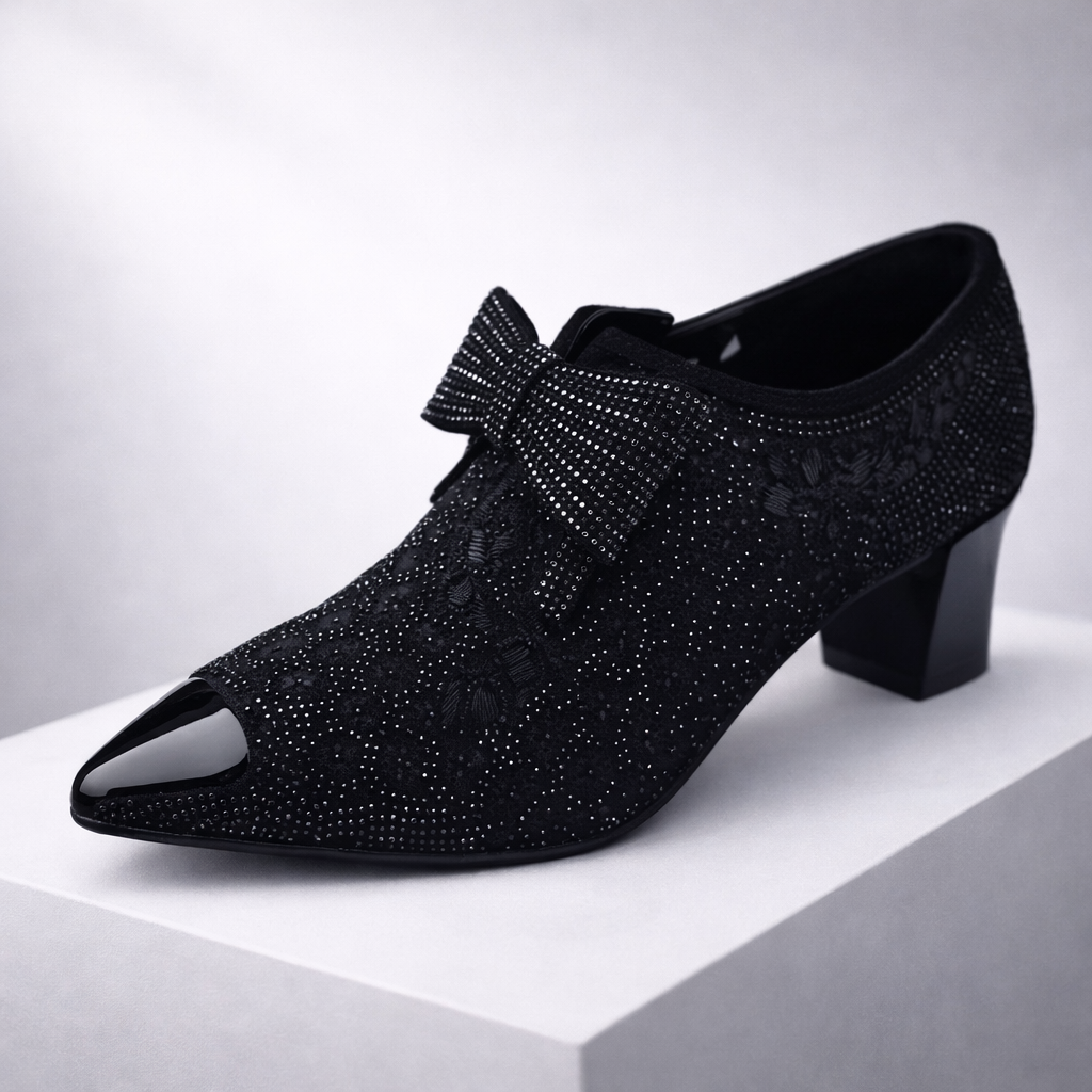 Black rhinestone-embellished pointed-toe block heel pump with bow detail and floral lace overlay on white display platform.