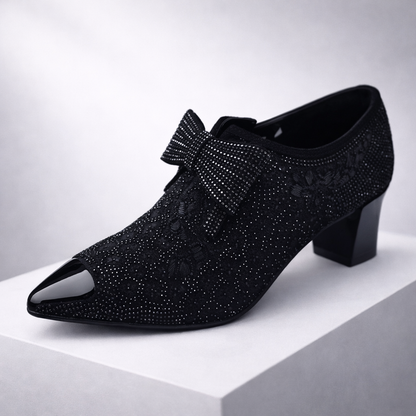 Black rhinestone-embellished pointed-toe block heel pump with bow detail and floral lace overlay on white display platform.