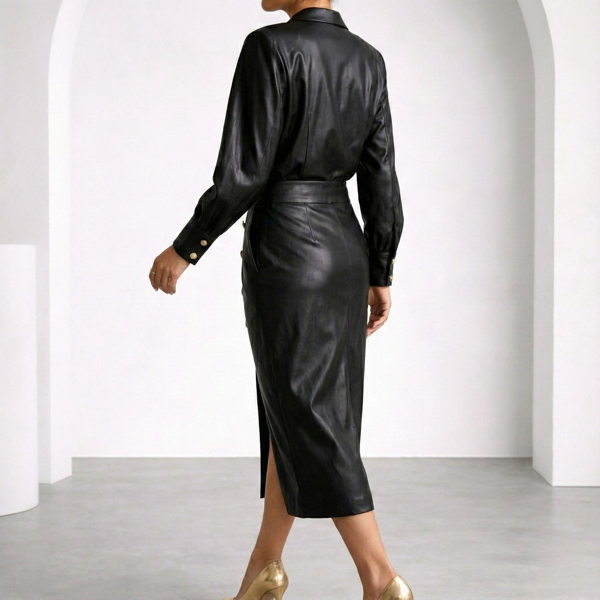 Woman wearing black faux leather belted midi dress with gold button cuffs and back slit, paired with gold heels.
