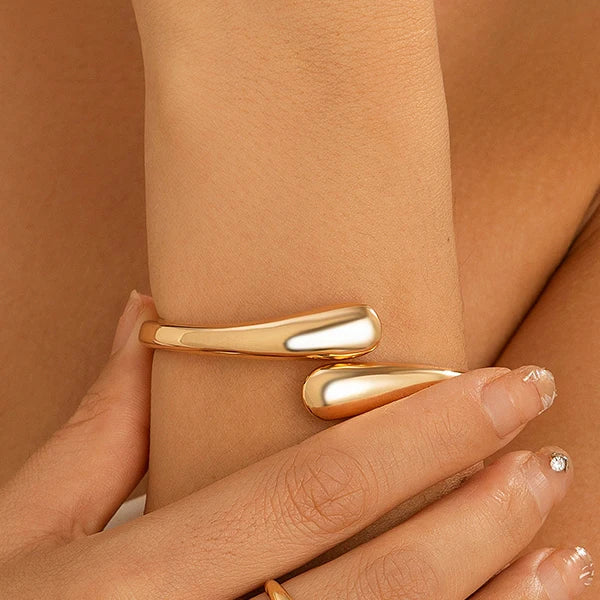 Gold tone open cuff bracelet worn on wrist, featuring sleek wrap design with tapered ends, minimalist modern style.