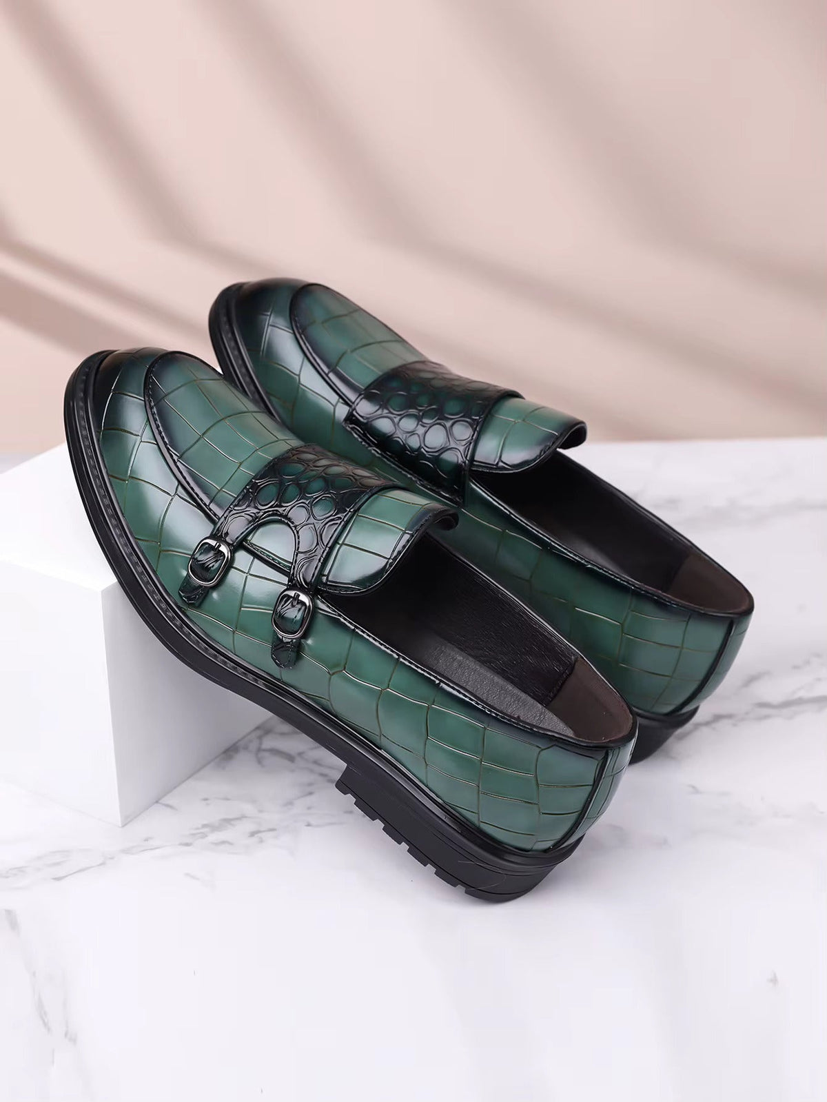 Green croc-embossed leather double monk strap loafers for men with black sole and silver buckles on marble surface.