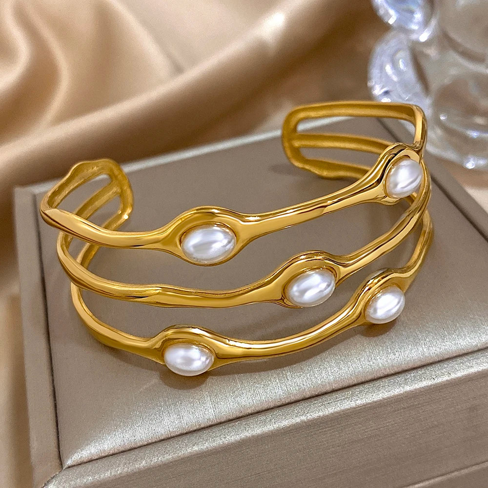 Gold tone open cuff bangle bracelet with three wavy rows and oval pearl accents, displayed on a jewelry box.
