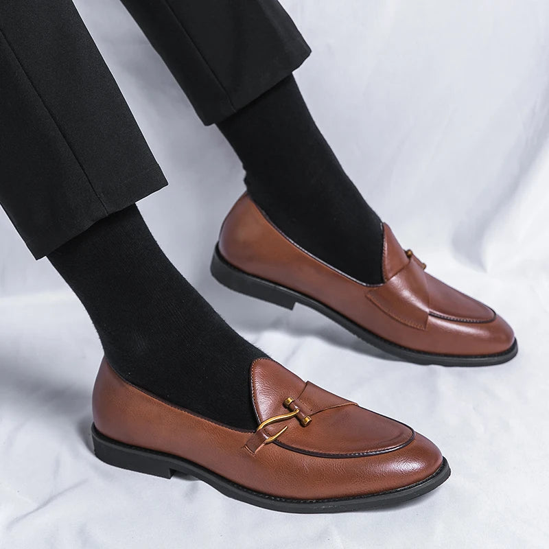 Brown leather men's slip-on loafers with gold metal buckle detail, worn with black socks and dress pants.