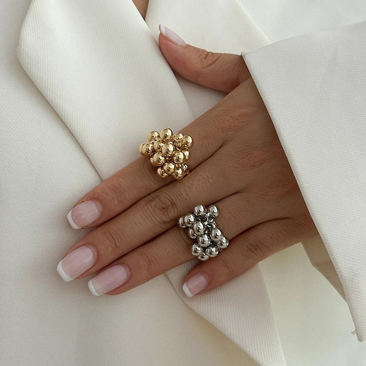 Hand wearing two adjustable beaded cluster rings in gold and silver, displayed against white fabric background.