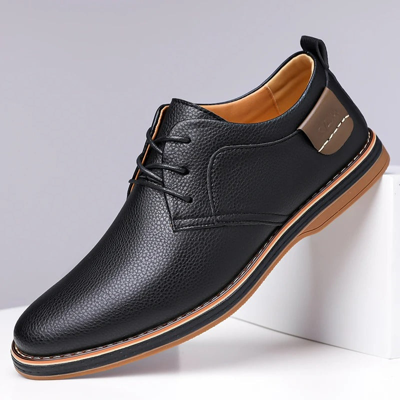 Black men's leather lace-up derby shoes with tan sole, pebbled texture, and brown heel accent on white display block.