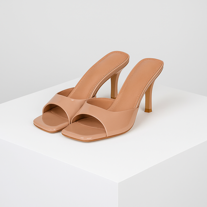 Nude patent leather square-toe mule heels with slim stiletto heel displayed on white pedestal