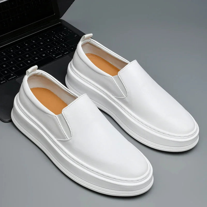 White leather slip-on loafers with thick white sole and tan insole, displayed next to a laptop on gray surface.
