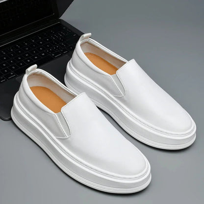 White leather slip-on loafers with thick white sole and tan insole, displayed next to a laptop on gray surface.