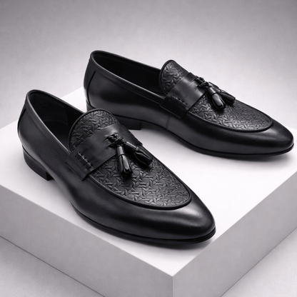 Black leather tassel loafer dress shoes for men with embossed toe cap and slip-on design, displayed on white platform.