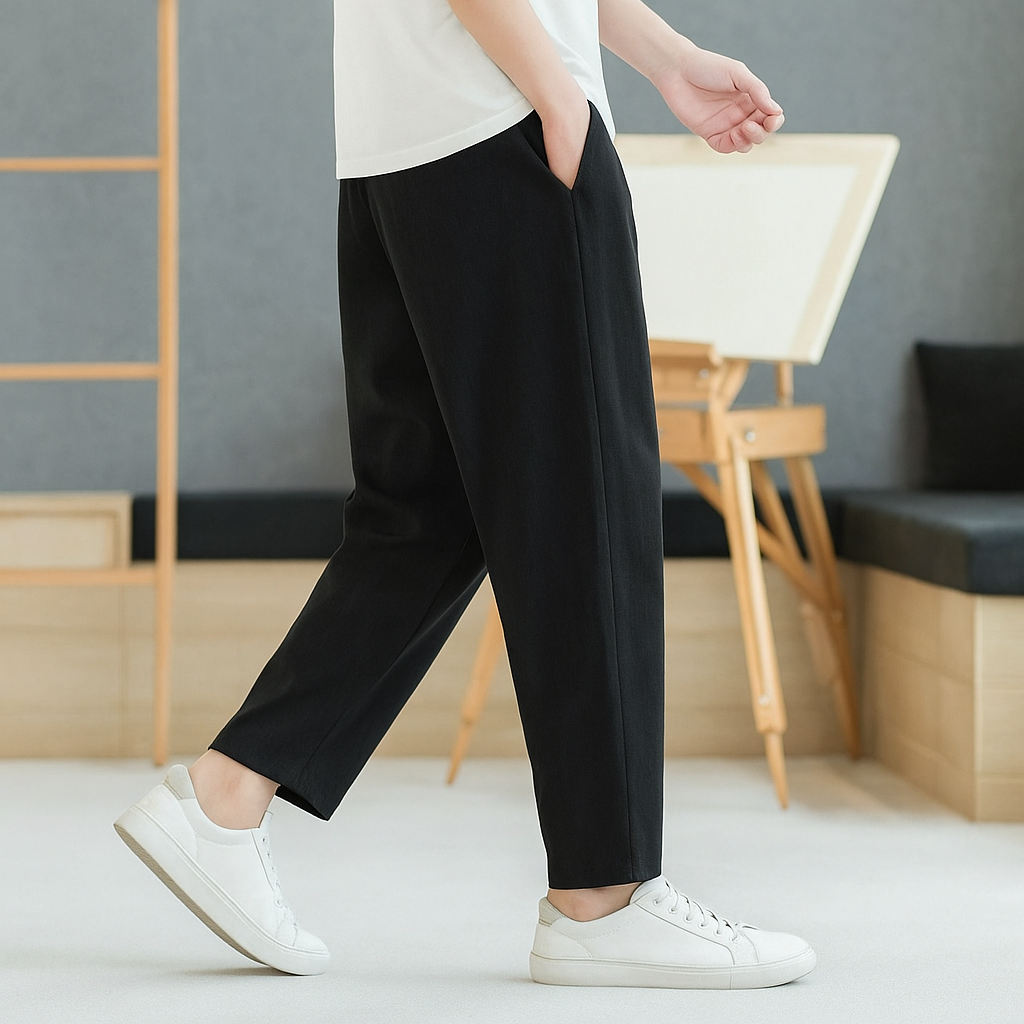Man wearing black tapered linen-cotton pants with white t-shirt and white sneakers, walking in minimalist indoor setting.