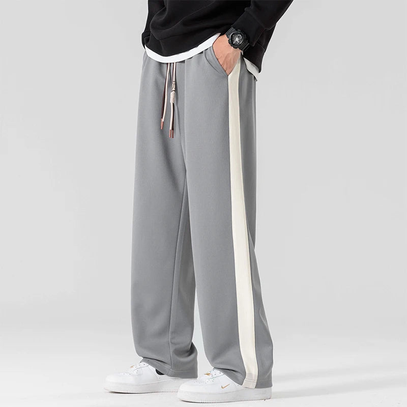 Gray wide-leg men's sweatpants with white side stripe, drawstring waist, styled with black sweatshirt and white sneakers.