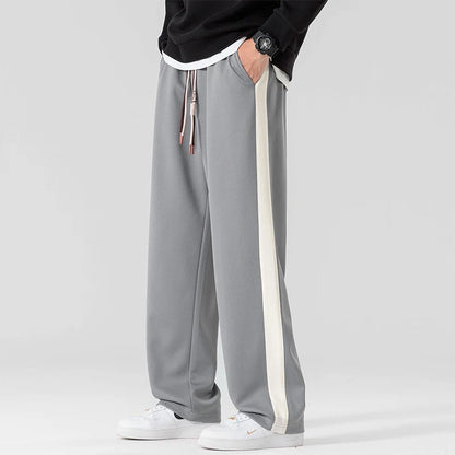 Gray wide-leg men's sweatpants with white side stripe, drawstring waist, styled with black sweatshirt and white sneakers.