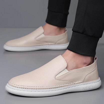Beige leather slip-on loafers with white sole and elastic gusset, worn with black jogger pants on gray background