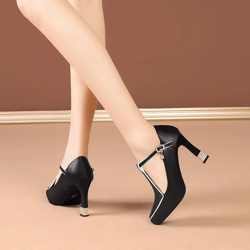 Black T-strap block heel pumps with gold buckle charm and white trim detailing, shown on crossed legs from behind.