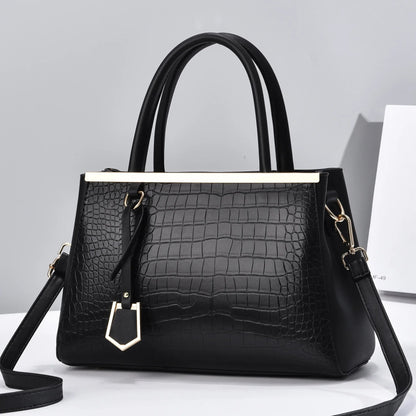 Black croc-embossed structured handbag with gold-tone bar detail, dual top handles, decorative charm, and removable crossbody strap.