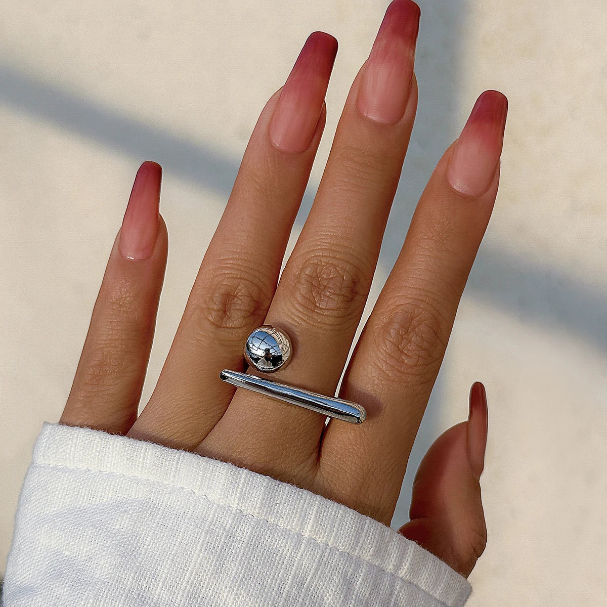 Hand wearing an adjustable silver bar and ball open ring with long ombre pink-to-red square nails and white sleeve.
