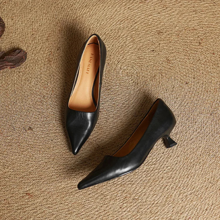 Black leather pointed-toe pumps with kitten heels and tan interior lining displayed on textured burlap surface.