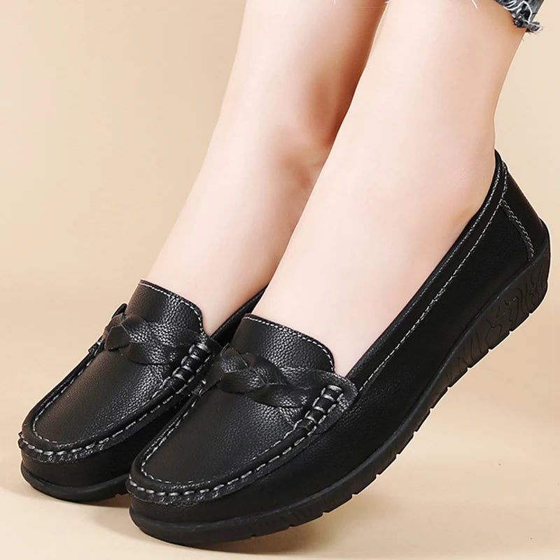 Black women's leather slip-on loafers with braided detail and white stitching, shown on model's feet against beige background.