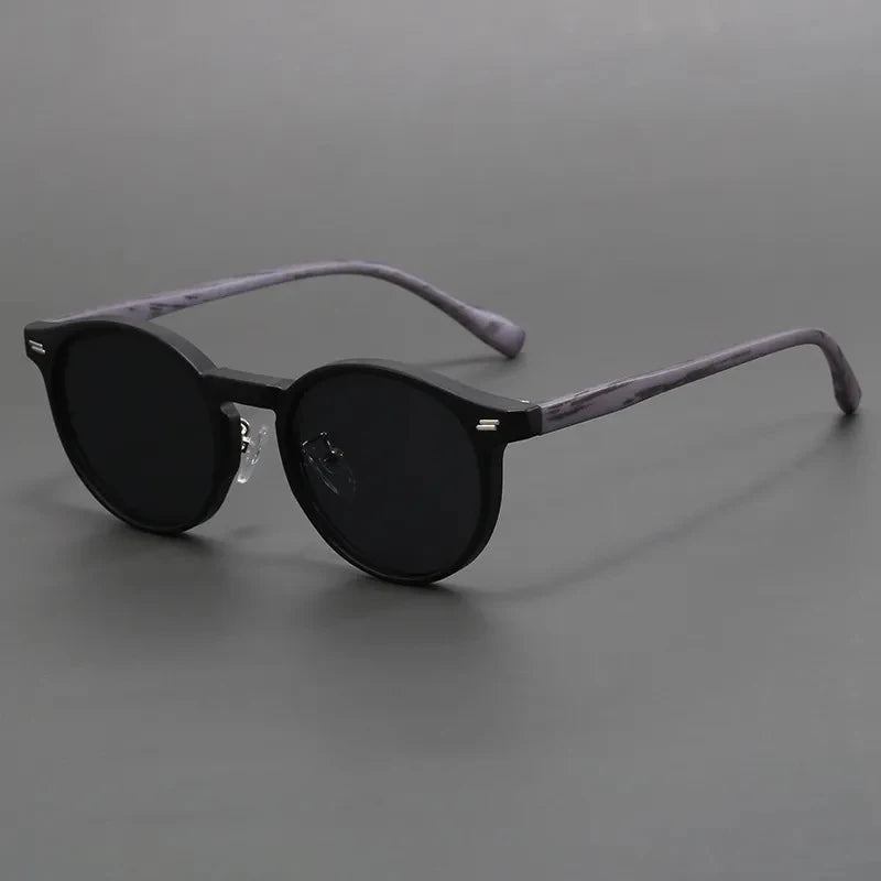Round black sunglasses with dark tinted lenses and marbled gray temple arms on a neutral gray background