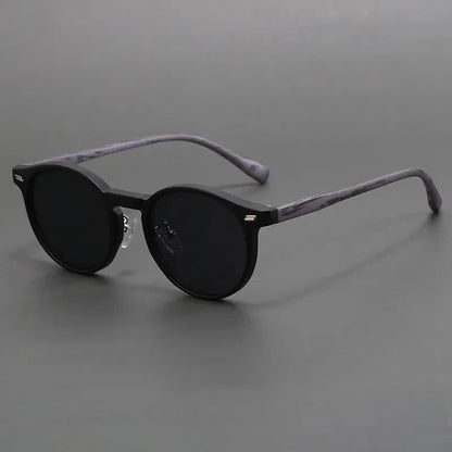Round black sunglasses with dark tinted lenses and marbled gray temple arms on a neutral gray background