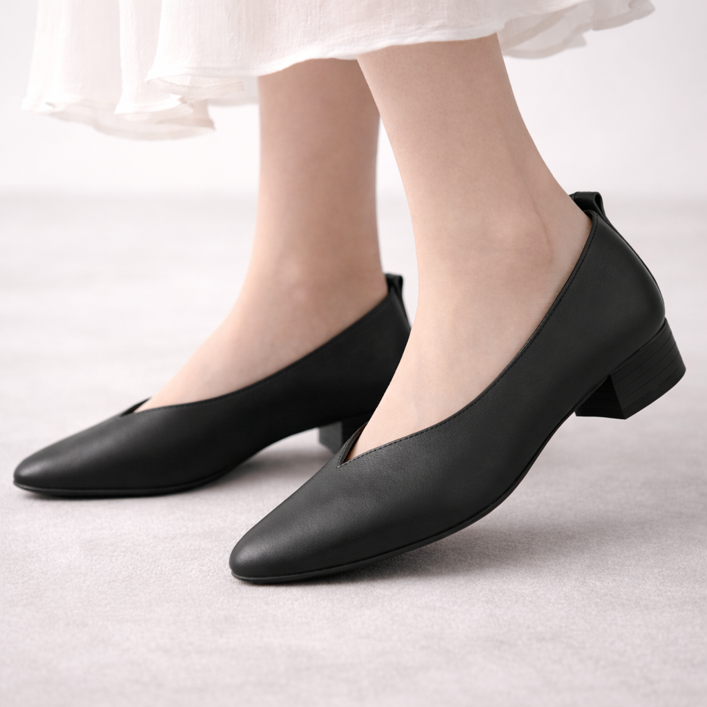 Black leather block heel pumps with V-cut vamp and pointed toe, worn by woman in white dress on grey background.