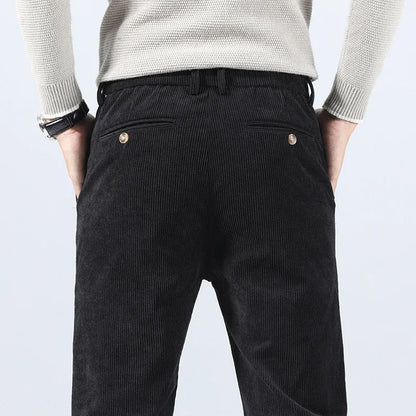 Back view of men's black corduroy slim fit pants with button back pockets, paired with a cream knit sweater.