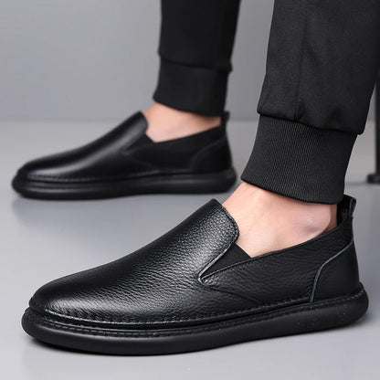 Black leather slip-on loafers for men with pebbled texture, elastic side gussets, and cushioned sole, styled with black joggers.