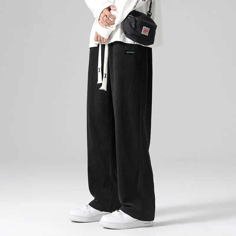 Black men's corduroy wide-leg sweatpants with drawstring waist, styled with white sneakers and crossbody bag.