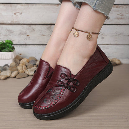 Women's burgundy leather slip-on loafers with decorative stitching and bow detail, shown with gold coin anklet.