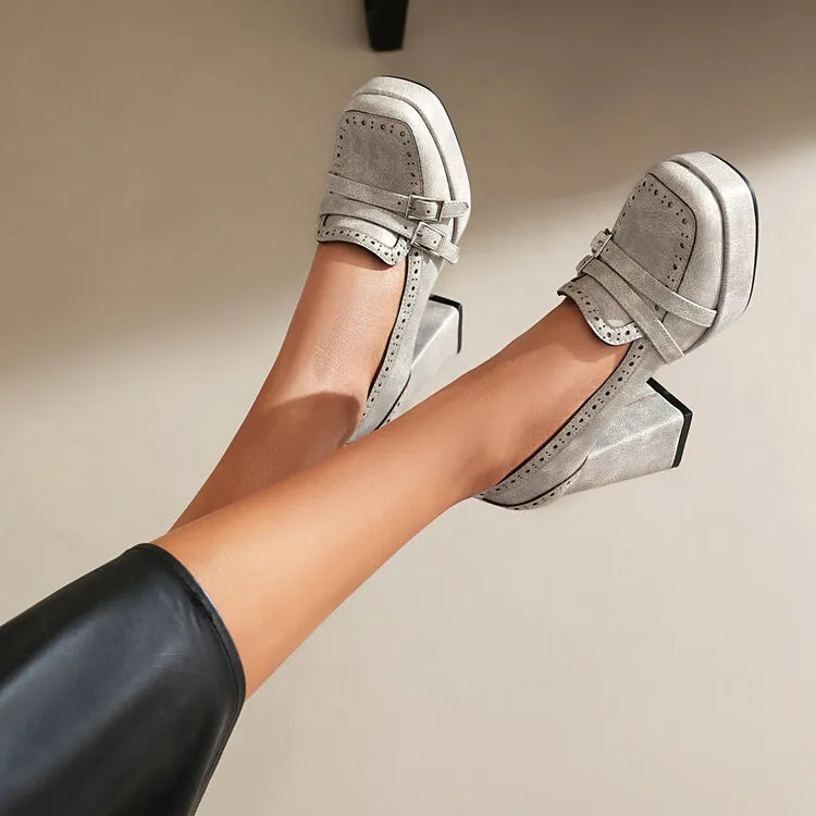 Women's gray suede platform block heel loafers with brogue detailing and buckle strap, shown on raised legs.