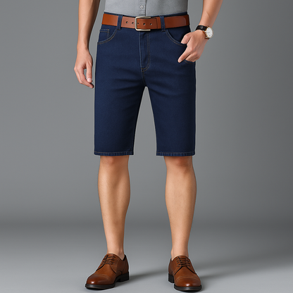 Men's navy blue stretch denim shorts with brown leather belt, paired with brown oxford shoes and gray shirt.