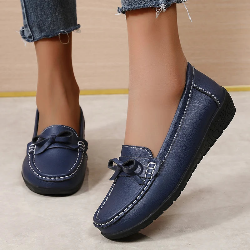 Navy blue leather women's slip-on loafers with bow detail and white stitching, paired with frayed denim jeans.