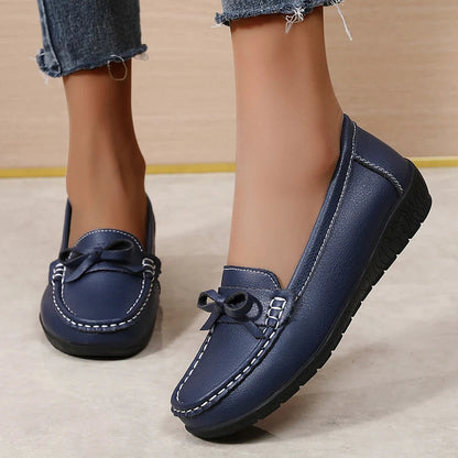 Navy blue leather women's slip-on loafers with bow detail and white stitching, paired with frayed denim jeans.
