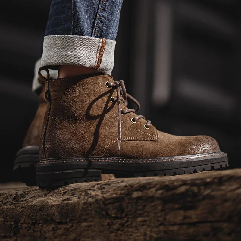 Men's brown suede lace-up ankle boots with rugged lug sole, styled with cuffed selvedge denim jeans on a wooden surface.